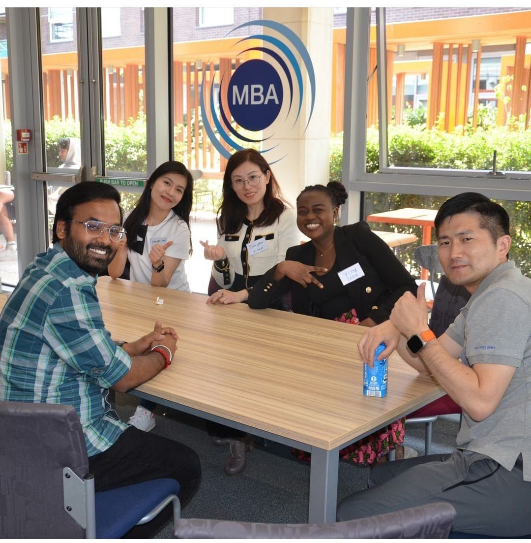 Five people sitting around a table in an MBA classroom, smiling and gesturing towards the camera.