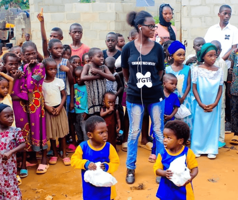 Group of children and adults standing together outside with some holding white bags, diverse clothing styles visible.